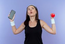 young-fitness-woman-headband-holding-smartphone-dumbbell-looking-confused-standing-blue-wall_141793-61159-2244002